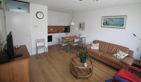 A living room with a brown leather sofa, a dining table with chairs, a kitchen area, and a clock on the wall.