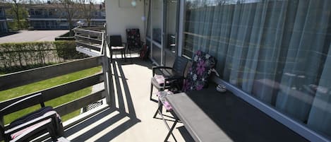 Un balcón con vistas a un jardín, una mesa con sillas y una ventana con cortinas.