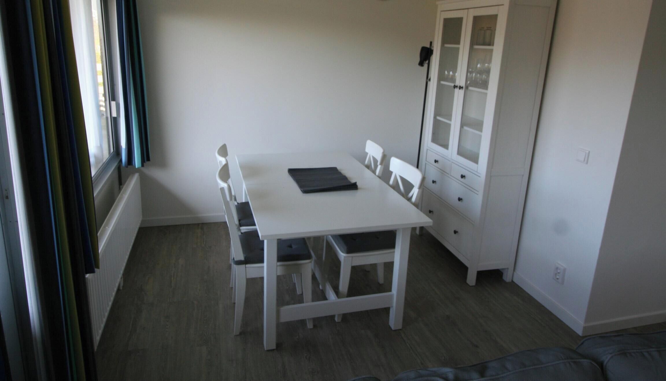 Una habitación pequeña y blanca con una mesa, sillas, un armario con puertas de cristal y una ventana con cortinas.