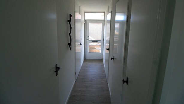 A narrow hallway with white walls and a wooden floor, leading to a door with a window.