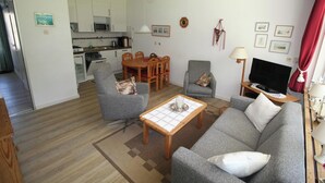 Un salón con un sofá gris, dos sillones, una mesa de centro y un televisor. Hay una zona de comedor con una mesa y sillas, y una cocina al fondo.