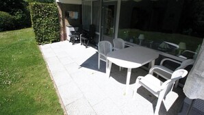 Un patio con una mesa y sillas, un seto y una puerta de vidrio que da a una zona cubierta.