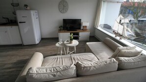 A modern living room with a sofa, a TV, and a window with a view of buildings. - 2 bedroom accommodation in Westkapelle (Westkapelle)