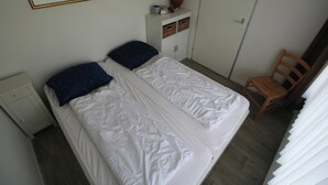Parivuode valkoisilla vuodevaatteilla ja kahdella tummansinisellä tyynyllä, pieni yöpöytä, puinen tuoli ja peilillä varustettu vaatekaappi.