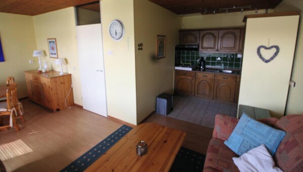 A living room with a wooden table, a couch with patterned cushions, a kitchen with wooden cabinets, and a clock on the wall. - 2 bedroom accommodation in Westkapelle (Westkapelle)