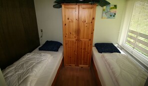 A small room with two single beds, a wooden wardrobe, and a window with blinds.