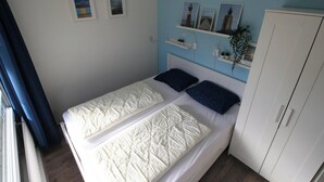 A single bed with white bedding, a headboard, and two pillows. There is a white wardrobe to the right and a window with a blue curtain on the left.