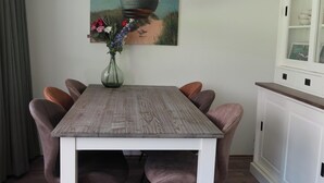 A dining table with chairs, a vase with flowers, a painting on the wall, and a cabinet with glassware.