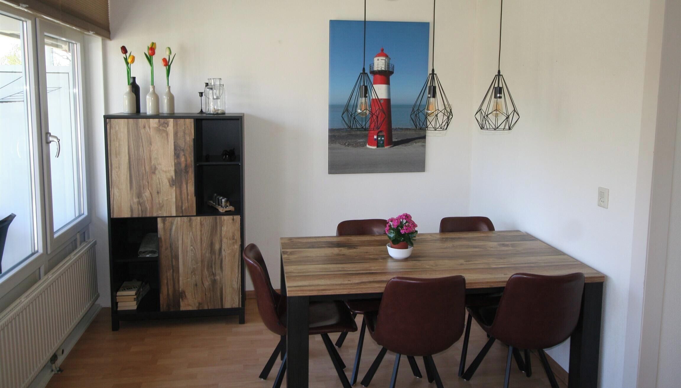 A dining area with a wooden table, four chairs, a vase with flowers, a painting of a lighthouse, and pendant lights.