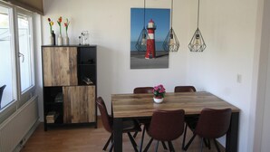 Un coin repas avec une table en bois, quatre chaises, un vase de fleurs, un tableau représentant un phare et des lampes suspendues.