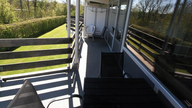 Un balcon donnant sur une pelouse et des arbres bien entretenus, avec une chaise et une petite table.