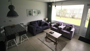 Ein Wohnzimmer mit einer Couch, zwei Sesseln, einem Couchtisch und einem Esstisch mit Stühlen. An der Wand hängen Bilder in Rahmen, eine Lampe steht dort, und es gibt einen Ausblick durch ein Fenster.