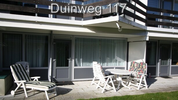 Ein modernes Gebäude mit Balkon, Außenbestuhlung und einem Schild mit der Aufschrift 'Duinweg 117'.