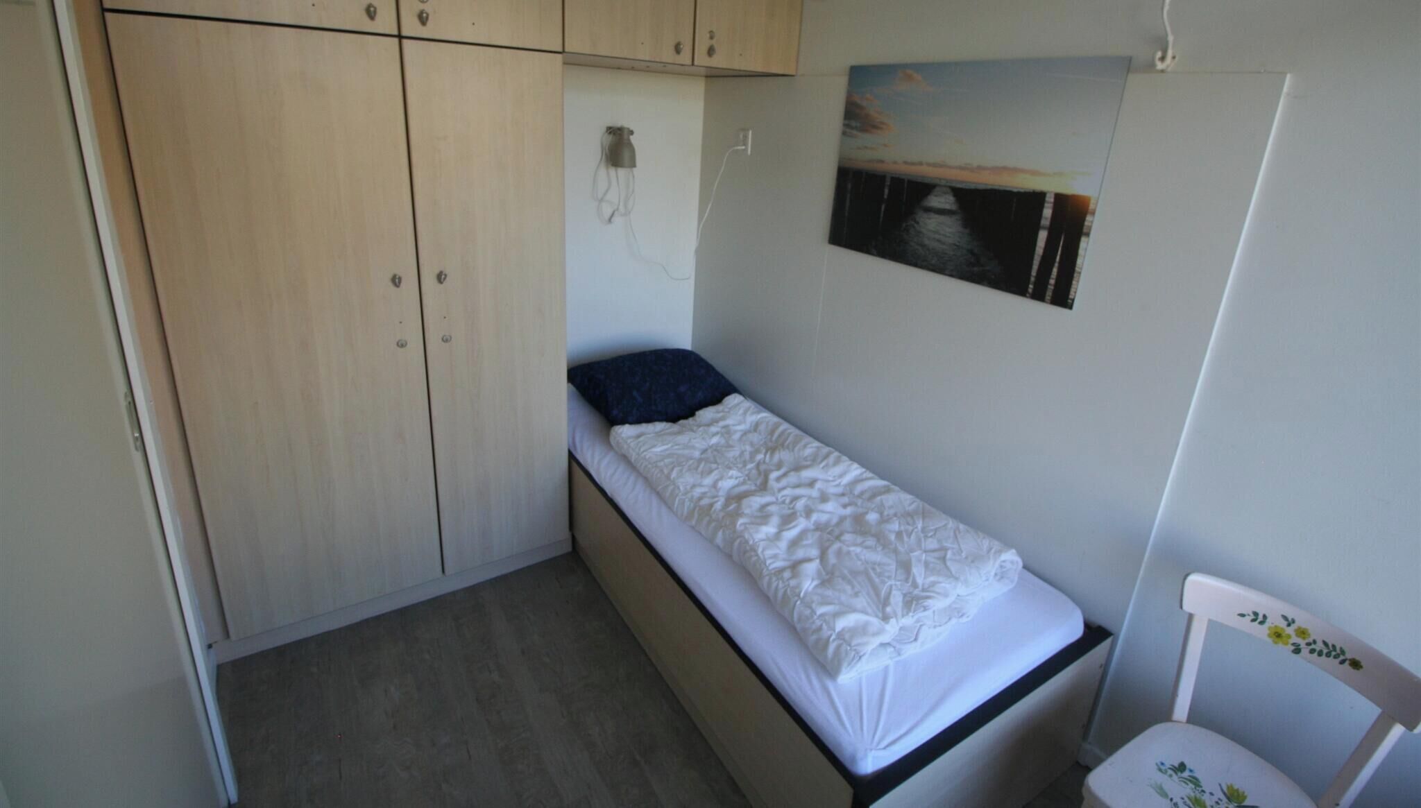 A small bedroom with a single bed, a wooden wardrobe, a chair, and a wall-mounted picture.