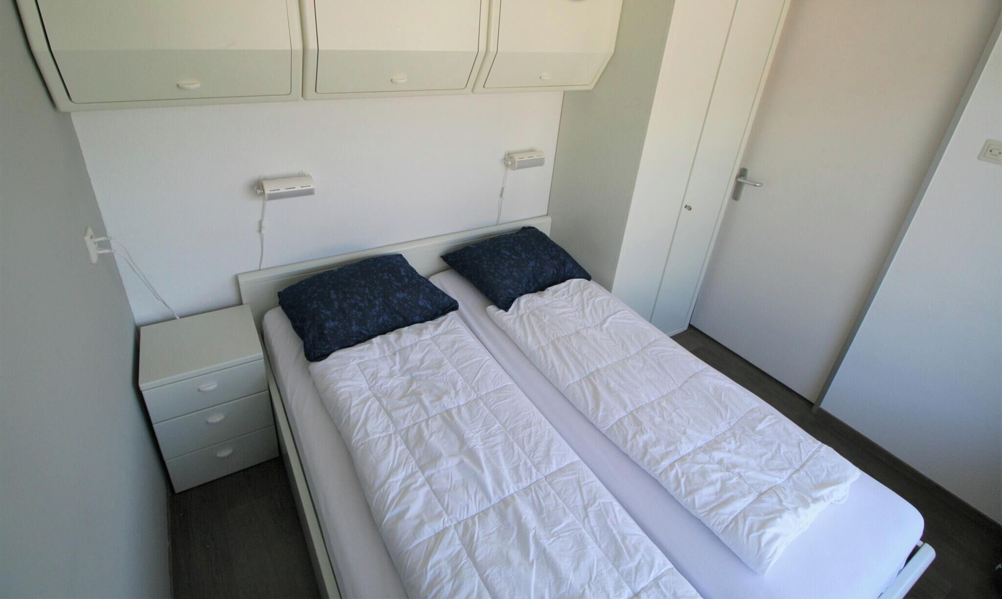 Un lit simple avec une couette blanche, deux oreillers, une table de chevet, une armoire et une porte.