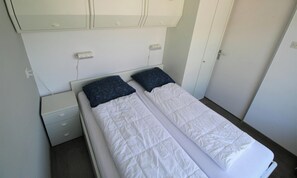A single bed with a white quilt, two pillows, a nightstand, a wardrobe, and a door.