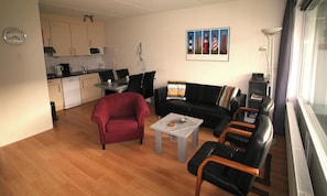 Uma sala de estar com uma poltrona vermelha, um sofá preto e uma mesa de centro. Cozinha visível ao fundo, com armários e um micro-ondas.