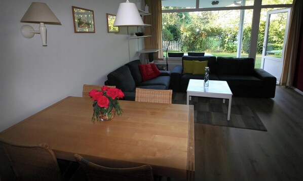 A living room with a wooden table, a sofa, a white coffee table, and a window with a view of greenery.