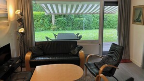 A room with a black leather sofa, a wooden coffee table, a TV, and a large window with a view of a garden and outdoor seating.