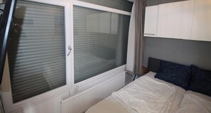 Ein Schlafzimmer mit Bett, Fenster mit Jalousien, Heizkörper und Kleiderschrank.