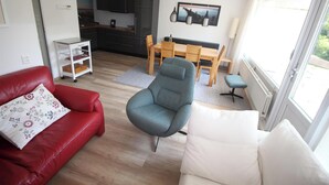 Un salon avec un canapé rouge, un fauteuil gris, un canapé blanc, une table à manger avec des chaises, et un coin cuisine à l’arrière-plan.