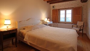 A bedroom with a large bed, a nightstand, a chair, a window, and wooden beams on the ceiling. - Charming authentic Norman house with fireplace and pets allowed (Barenton)