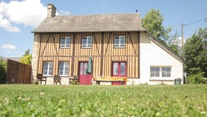 A two-story house with a sloped roof, a red door, and a small garden in front. - Charming authentic Norman house with fireplace and pets allowed (Barenton)