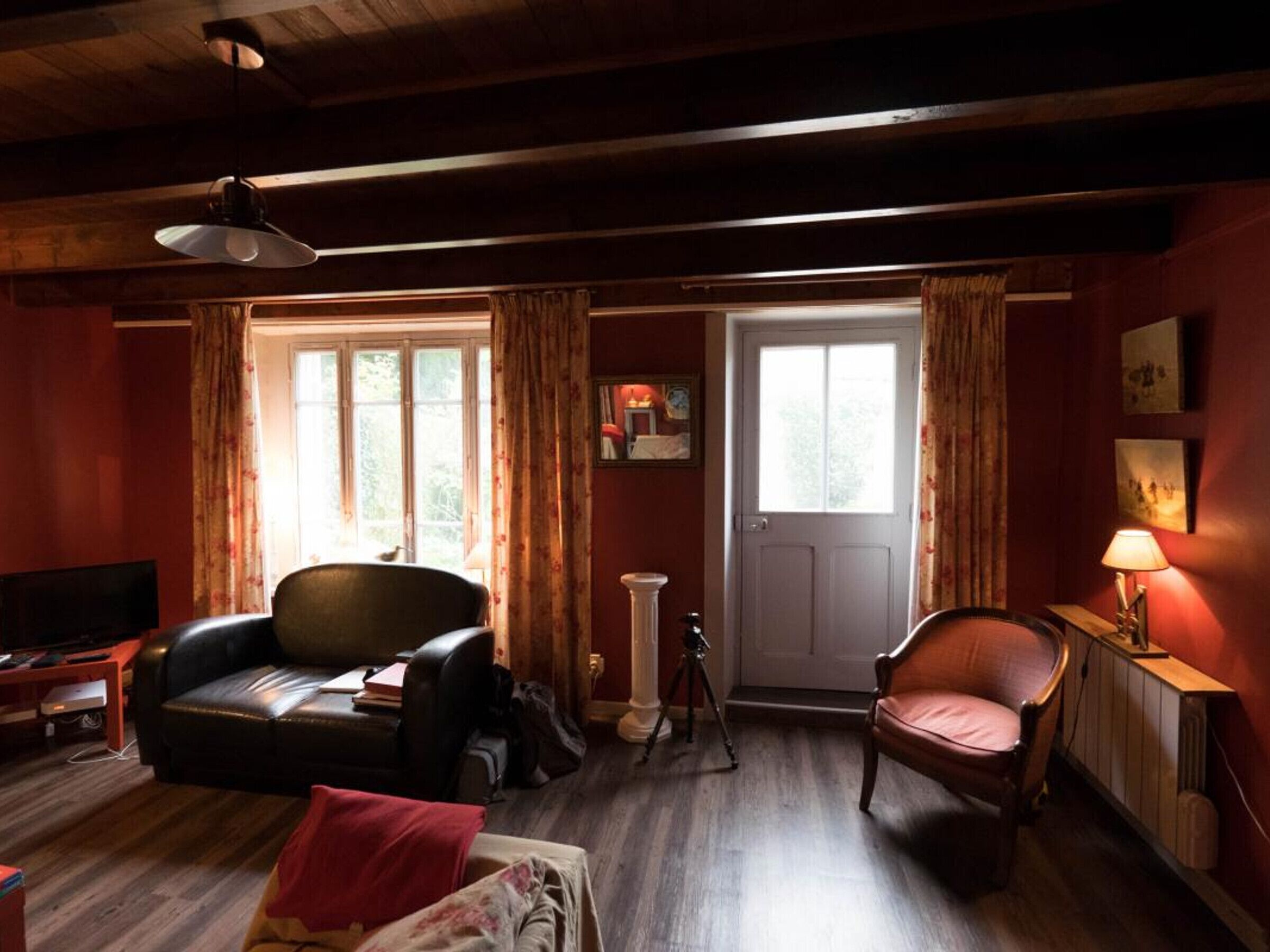 Un salon chaleureux avec un plafond en bois, un canapé en cuir noir, un fauteuil en bois, une petite table avec une lampe et une porte donnant sur l’extérieur.