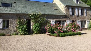 A stone building with a green roof, a gravel driveway, and pink flowers in the foreground. - Cozy house with garden, pets allowed, WiFi in La Hague (La Hague)