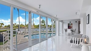 A modern hotel room with large sliding glass doors to a pool area, a dining area with a table and chairs, and a kitchen visible through an open door. -  Villa NOVA (North Bay Village)