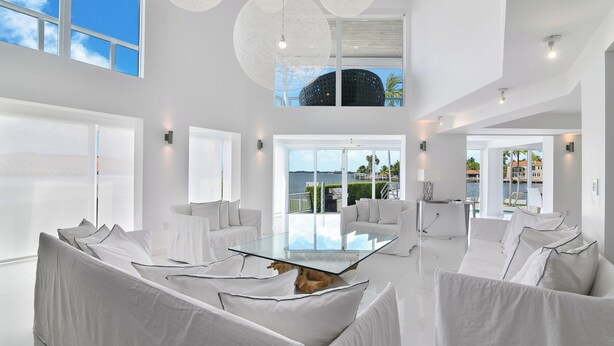 A spacious living area with a glass coffee table, white sofas, and a view of a body of water through large windows. -  Villa NOVA (North Bay Village)