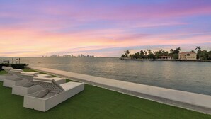 A waterfront property with a row of white lounge chairs on a grassy area, a concrete pathway, and a view of a city skyline across the water. -  Villa NOVA (North Bay Village)