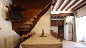 A dining area with a table set for six, wooden chairs, a wooden staircase, a kitchen visible through an open door, and a television mounted on the wall.