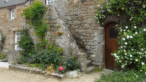 A stone building with a wooden door and windows, surrounded by greenery and flowers.