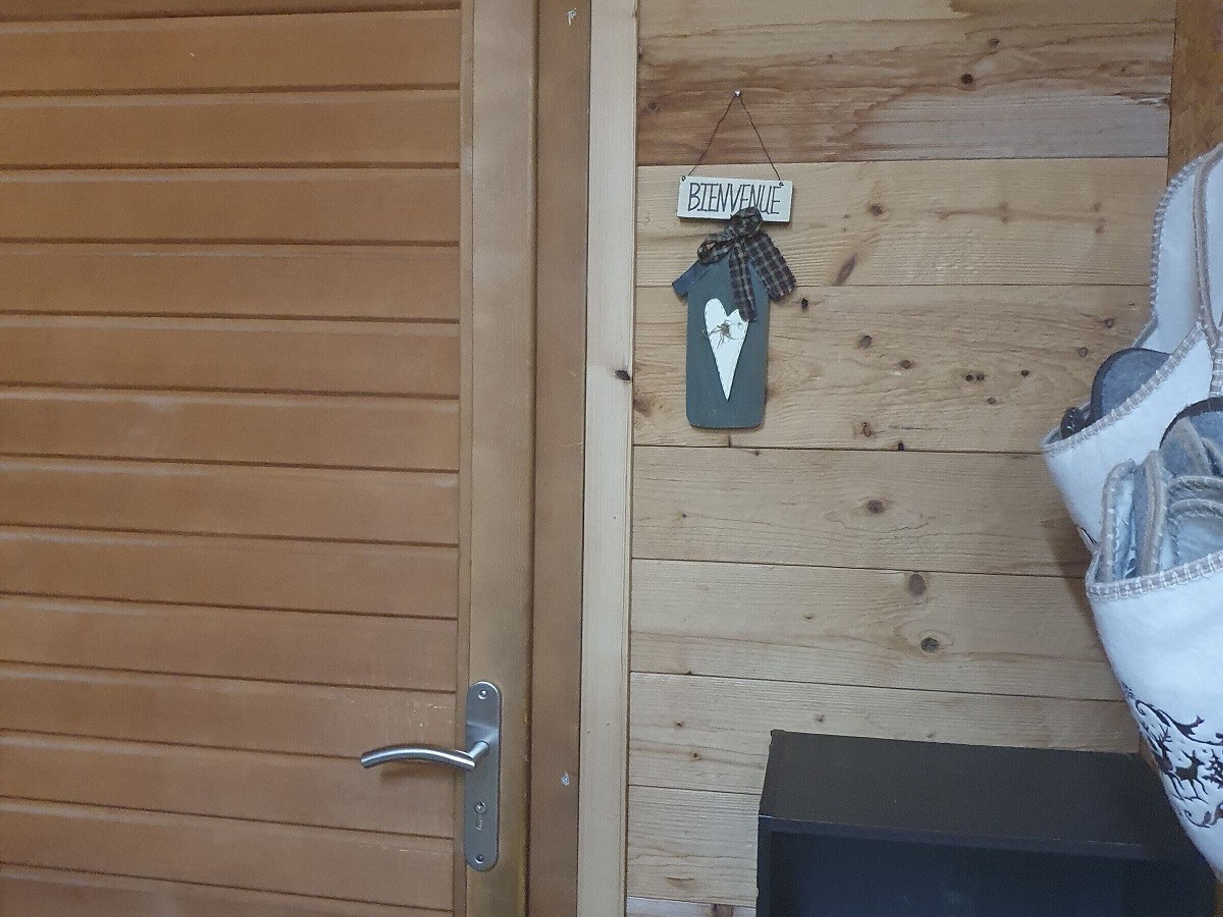 Een houten deur met een metalen handgreep, een houten wand met een 'BIENVENUE' bord, en een klein decoratief object dat aan de wand hangt.