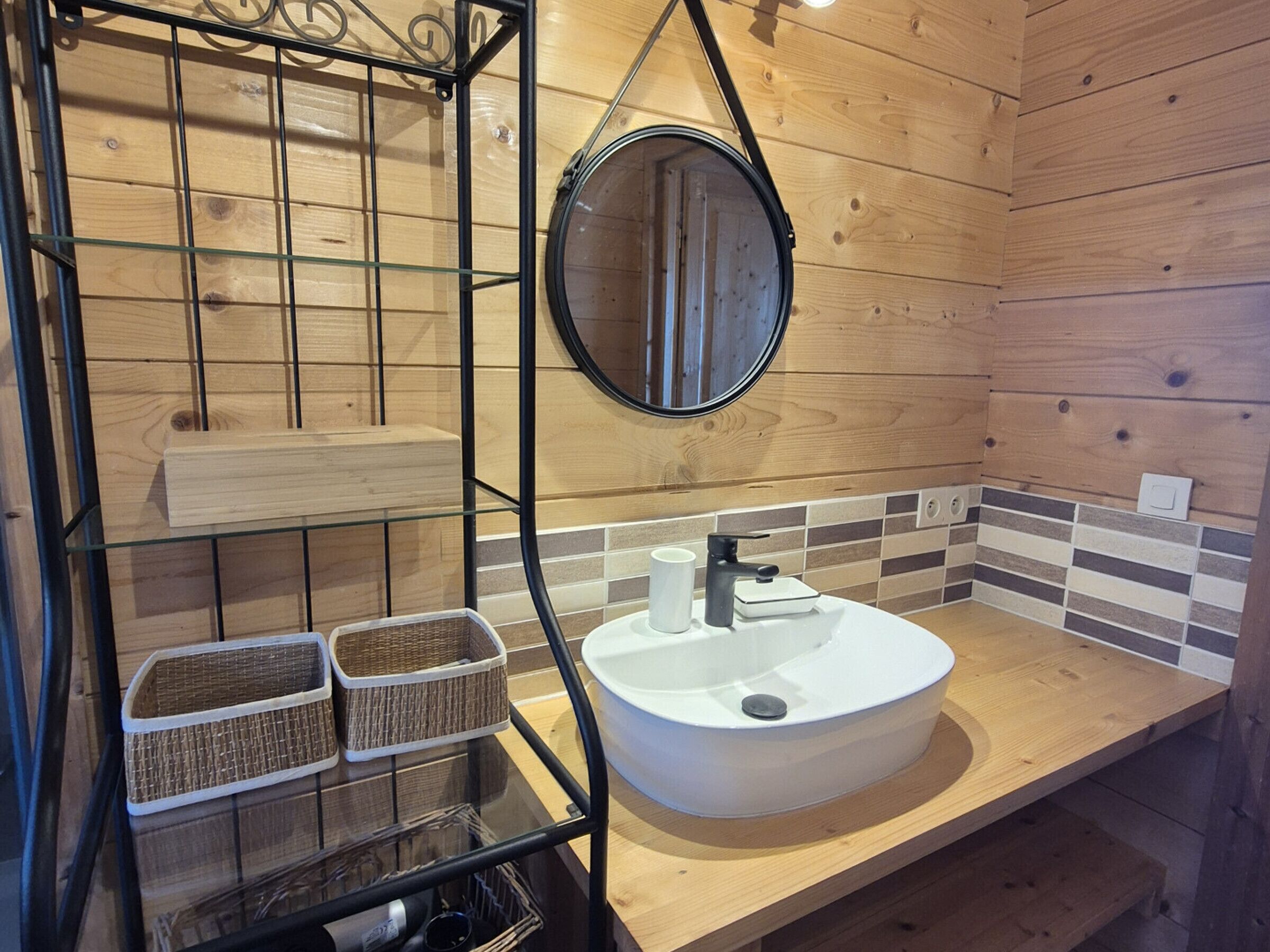 A bathroom with a wooden interior, a round mirror, a white sink, and wicker baskets on a glass shelf.