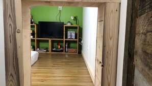 Un étroit couloir en bois mène à une pièce dont un mur est peint en vert, qui contient une télévision et des étagères remplies de divers objets.