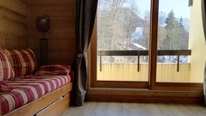 Ein gemütliches Zimmer mit einer Holzcouch, einem Standventilator und einem großen Fenster mit Blick auf Bäume und Gebäude.