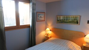Ein Schlafzimmer mit einem Bett, zwei Nachttischlampen, einem mit Vorhängen versehenen Fenster, einem gerahmten Bild an der Wand und einem Gemälde über dem Bett.