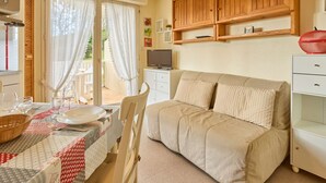 A cozy living room with a beige sofa, a dining table set with plates and glasses, a television on a stand, and wooden shelves above the sofa. - Charming T2 in Barbotan: WIFI, Pets, Parking (Barbotan-les-Thermes)