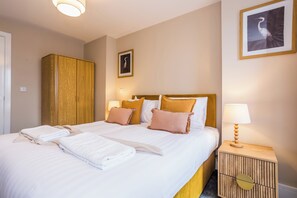 A neatly made double bed with white and beige bedding, a wooden wardrobe, a nightstand with a lamp, and a framed picture on the wall.