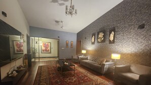 A spacious living room with a large sofa, a coffee table, and a patterned rug. There are two wall-mounted lamps and a chandelier.