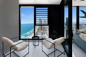 Una habitación moderna con un gran ventanal con vista a la playa y al paisaje urbano, dos sillas blancas y una mesita redonda.