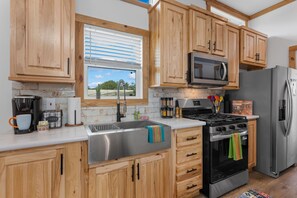 A kitchen with wooden cabinets, a stainless steel sink, a microwave, a stove, and a refrigerator. - Rancho Relaxo - On the 290 Wine Trail (Fredericksburg)