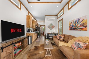 A modern living room with a large flat-screen TV displaying 'NETFLIX,' a wooden coffee table, a brown leather sofa, and a kitchen area with wooden cabinets and a window. - Rancho Relaxo - On the 290 Wine Trail (Fredericksburg)
