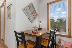 Uma área de refeições com uma mesa de madeira, cadeiras, um painel decorativo fixado na parede, uma janela com persianas e uma planta pendente.