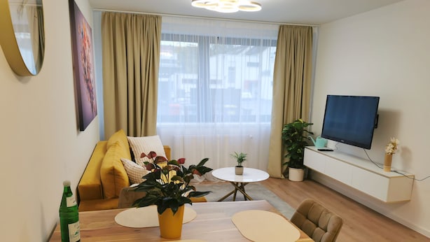 Moderni olohuone, jossa on puinen ruokapöytä, beigen värinen sohva ja seinälle asennettu taulutelevisio.
