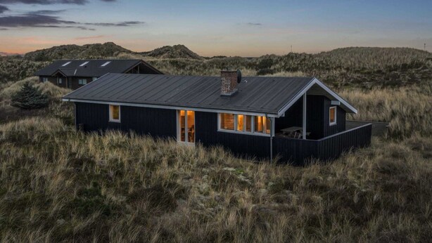 A modern house with a dark exterior, large windows, and a flat roof, surrounded by tall grass and dunes.