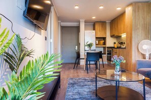 Un espace de vie moderne avec un téléviseur à écran plat fixé au mur, un coin repas avec des chaises, une cuisine avec des meubles en bois et un ventilateur sur le côté droit.