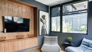Ein modernes Wohnzimmer mit einem großen Flachbildfernseher, einem Sitzsack, einer Couch und einem Fenster mit Blick auf Grünflächen.